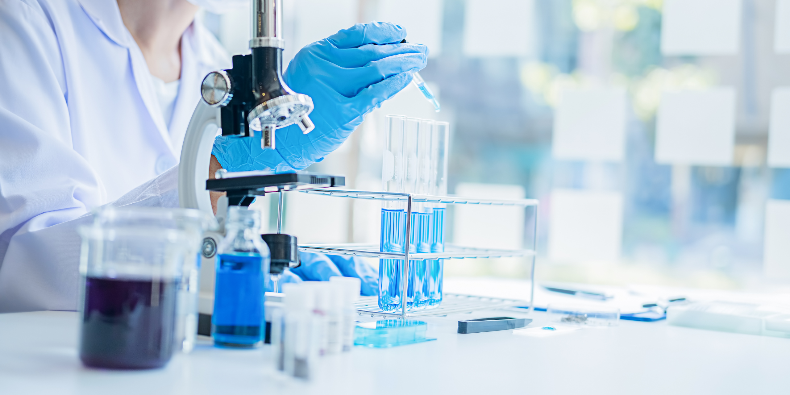 Scientific Research A researcher placing a liquid sample into a test tube, with a beaker of other liquid and a microscope nearby.